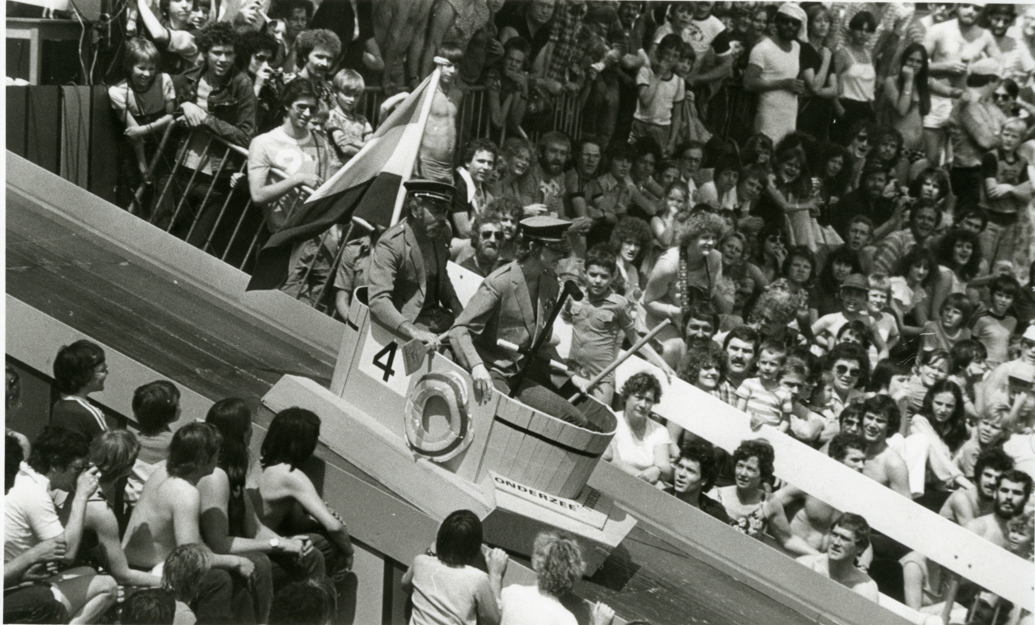 Zwart-wit foto uit 1979 van tobbedansen evenement. Veel toeschouwers en ronde tobbe met vlag en startnummer
